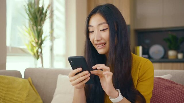 Smiling asian young woman use phone sitting on sofa at home. Social media text email enjoy distant communication. Social media. Close up. Slow motion