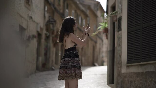 Side View Of A Young Girl In Backless Dress Taking Pictures Outside The Old House Using A Smartphone In The Street Of Mallorca, Spain.  - wide shot