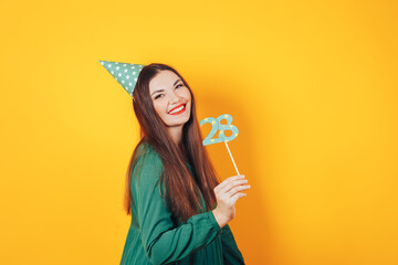 Beautiful woman celebrates her birthday on a yellow background in green dress and on her head festive cap holds the number 28 in her hand.