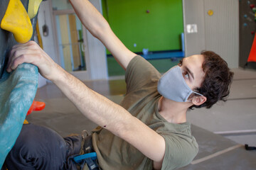 Young man wearing a COVID-19 pandemic mask climbs a bouldering wall in a climbing gym following social distance guidelines for exercise