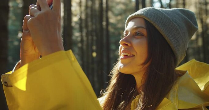 Close up portrait of happy Caucasian woman making photos on smartphone while standing in forest between trees. Beautiful female in woods taking pictures and tapping on cellphone. Tourism concept