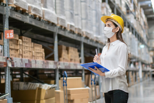 Female construction worker wearing protective face mask writing on clipboard checking product on aisle in warehouse factory. industrial social responsibility prevention Coronavirus. new normal working