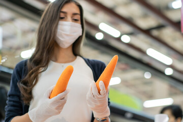 Woman wearing protective face mask and rubber glove holding carrot at vegetable grocery department store. shopping at supermarket in new normal lifestyle concept during Coronavirus or Covid pandemic.