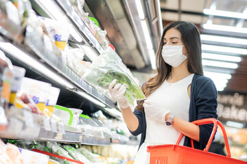 Beautiful woman wearing medical face mask and rubber glove holding grocery basket picking up vegetable on product shelf. shopping at supermarket in new normal lifestyle concept.