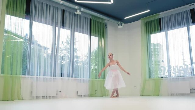 little girl in long skirt dances classical ballet doing jumps at windows with curtains in light studio slow motion