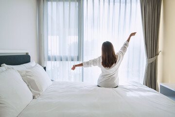 Back view of beautiful young Asian girl in white pajamas stretching after wake up in the morning on white bed in bedroom.