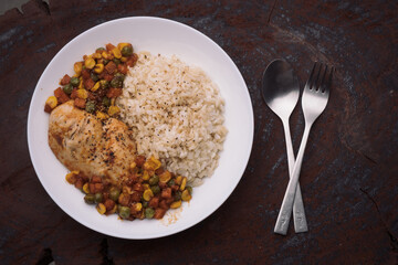 a dish of homemade roasted chicken breast and dice carrots, beans, corns with brown rice on dark red wooden table and cutlery