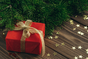 Christmas card with a red gift with a gold ribbon, fir branches and stars confetti on a wooden background. Copy space.
