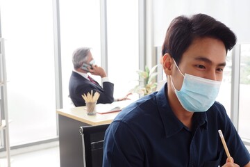 Young Asian male wearing face mask working in the office to prevent the Covid 19 virus for good health and hygiene at workplace and social distancing concept