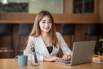 Young pretty asian businesswoman working on laptop while sitting at the table in office. Looking at camera.