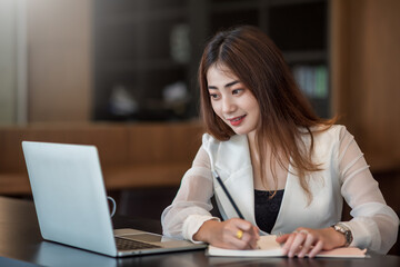 Charming asian businesswoman working on laptop and taking note while sitting at the table in office.