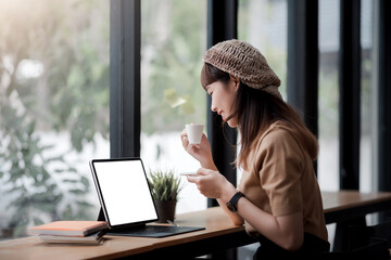 Mockup. Image of asian woman drinking coffee and working on a laptop with a blank white screen in a...