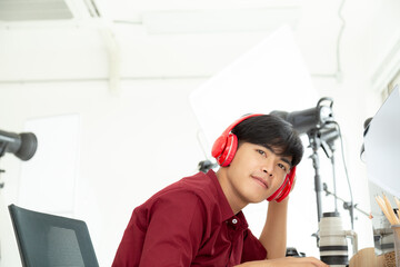 Portrait young happy asian photographer in red shirt working and listening music from red modern head phone in his studio.