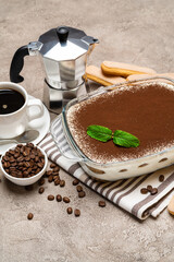 Traditional Italian Tiramisu dessert in glass baking dish, cup of espresso and mocha coffee maker on concrete background