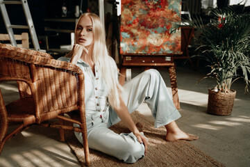 Beautiful blond dressed in styled clothes relaxing on carpet leaning on armchair around stylish bamboo furniture in warm hotel room.