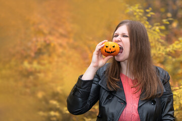 A beautiful girl bites a pumpkin on the background of an autumn forest. A woman with a pumpkin in a black leather jacket and pink jacket. Halloween.