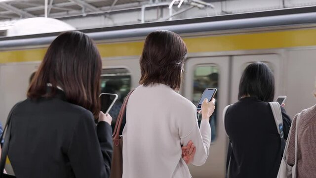 電車待ちでスマホを操作する若い女性たちの後姿