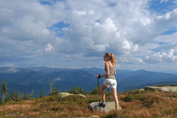 Naklejka premium Young woman hiking up rock with sticks against a blue sky with clouds. A woman climbing on top of mountains.