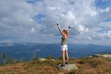 Young woman hiking up rock with sticks against a blue sky with clouds. A woman climbing on top of mountains.