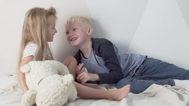 The older brother kisses his younger sister on the cheek. Children play at home in the morning with a teddy bear