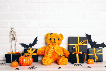 Halloween holiday background with orange toy bear, gifts and decorations against a white brick wall.