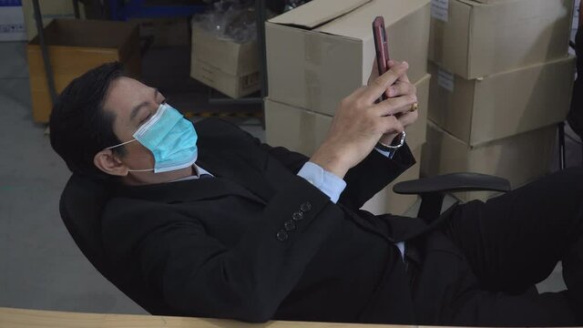 Lazy asian warehouse manager wearing face protective medical mask lying on chair using mobile phone   instead of working at storehouse