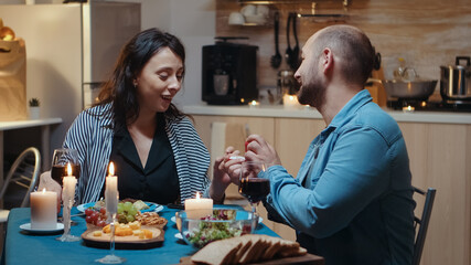 Obraz na płótnie Canvas Young cheerful woman being surprised by marriage proposal of her husband. Man making proposal to his girlfriend in the kitchen during romantic dinner. Happy caucasian woman smiling being speechless