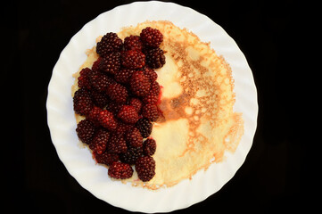 Ruddy pancake with ripe blackberries on white glass plate on a black table