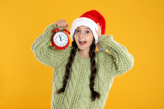 Girl in Santa hat with alarm clock on yellow background. Christmas countdown