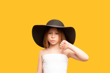 Skeptic child. Dislike gesture. Wrong choice. Bad idea. Portrait of disappointed spoiled young girl in black elegant hat rejecting offer showing thumb down isolated on orange copy space background.