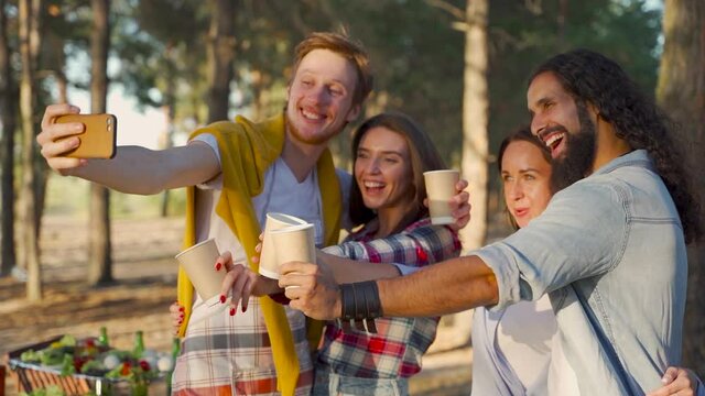 Group of young women and men taking a selfie with the phone outdoors. People enjoying a picnic in nature.