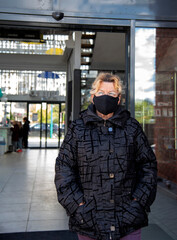 An elderly woman in a black jacket and a black medical mask walks out of the door of a shopping center.