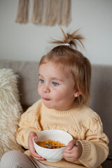 A cute little girl 2 years old in a cozy knitted sweater is having breakfast on the couch. Cozy. Fall.