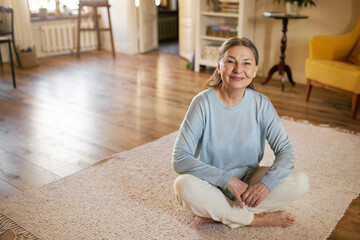 Indoor image of joyful happy middle aged woman wearing casual clothes sitting barefooted on white...