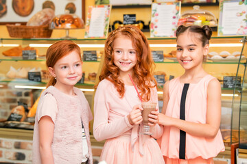 joyful children stand in a cafe against the background of showcase with sweets. A red-haired girl holds a mug of hot chocolate.