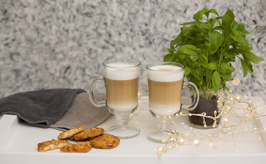 Two glass cups with coffee hot chocolate cappuccino latte with cookies on a white tray. Background in white and gray tones. Garland and bokeh