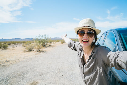 asian woman by car having jolly laugh is taking selfie with wild nature in desert region. female visitor taking photo background open land with clumps of bush and azure sky. summer grand tour concept