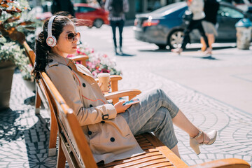 confident asian chinese woman in sunglasses listening to music from smartphone with headphones in city street. young girl with headset enjoy sunshine sitting on wood bench holding mobile phone.