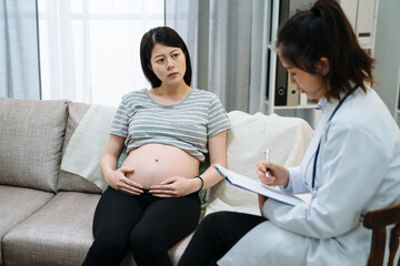 young asian korean pregnant woman sitting on sofa during psychotherapy. Worried maternity lady listening to psychologist counselor during therapy on couch at home. unhappy future mom with bare belly. - obrazy, fototapety, plakaty