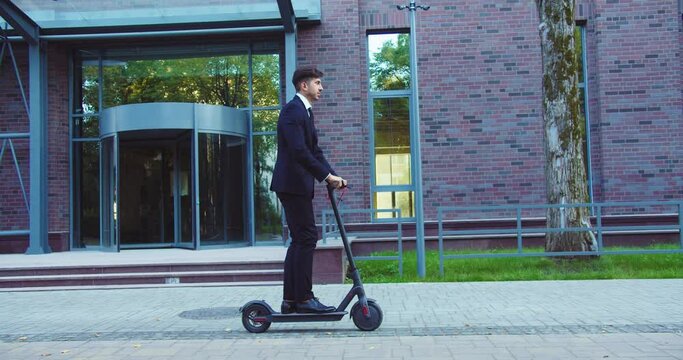 Good-looking Caucasian businessman riding electric kick scooter near modern office building. Successful adult male entreprenuer goinging to work by ecological transport. Wellness, success, job.