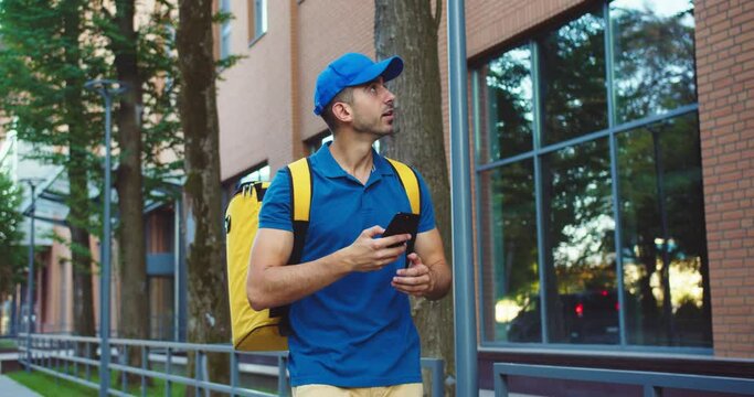 Attractive young male courier delivering food to offices in business district. Caucasian man worker with backpack using smartphone to find adress. App, service, technology, Internet concept.