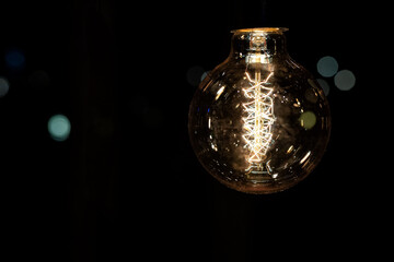 Glowing vintage old one tungsten lightbulb hanged on ceiling against black wall bokeh background....