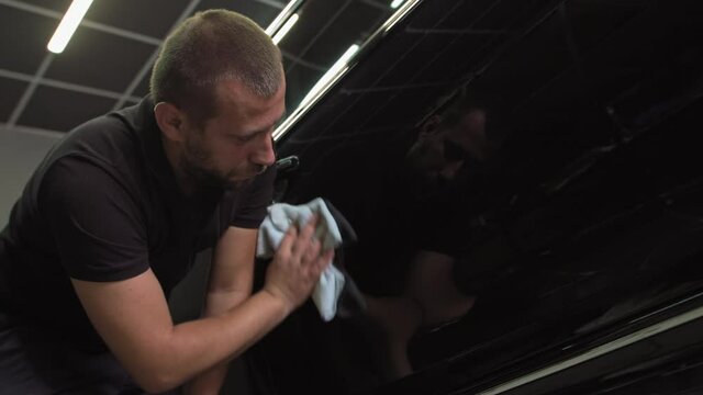 A male car mechanic wipes a black car with microfiber after restoring the body. Polishing car parts