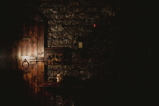 Mystical Dark Interior Of Medieval Room With Large Wooden Door And Skull On Table Against An Ancient Stone Wall. Amazing Background For Halloween Holiday. Copy Space, Text Place