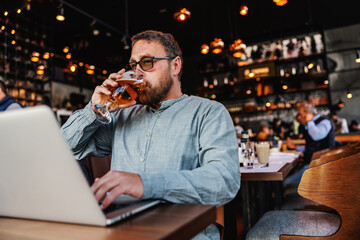 Middle aged bearded freelancer with eyeglasses sitting in a bar, drinking fresh cold light beer and...