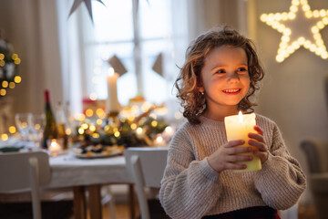Cheerful small girl standing indoors at Christmas, holding candle.