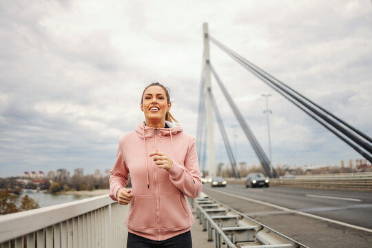 Smiling attractive fit sportswoman in tracksuit jogging on bridge on cloudy weather. Healthy lifestyle concept.