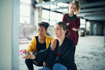 Group of teenagers girl gang indoors in abandoned building, using spray paint on wall.