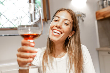Cute young caucasian blonde woman toasting with red wine glass looking at the camera at home - New...