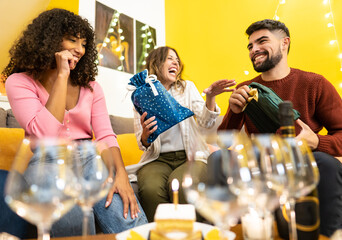 Two young beautiful caucasian friends give their gift to their black friend celebrating her birthday at home laughing and joking - Mixed race home party - Selective focus on curly woman to the left